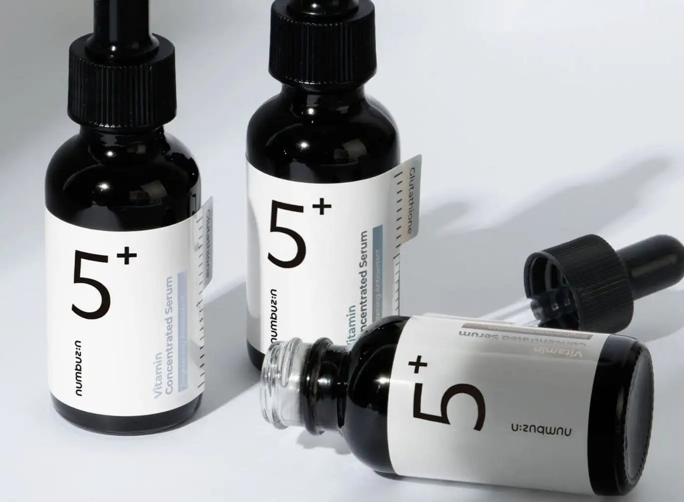 Three sleek black glass bottles with minimalist white labels and dropper caps, each prominently displaying the ’5+’ logo for numbuzn vitamin concentrated serum.