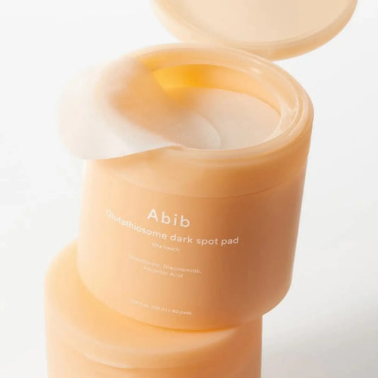 A peach-colored cylindrical container of abib glutathione dark spot pad holding translucent pads for brightening vitamin c toner use